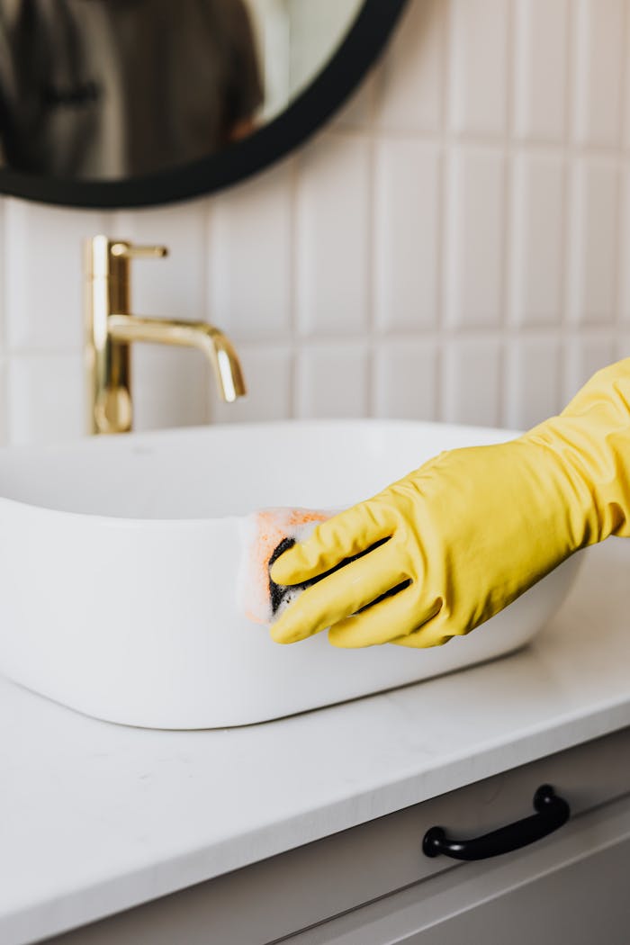 services-03 Crop faceless person in yellow rubber protective glove wiping surface of white sink in bathroom with sponge and cleaning agent
